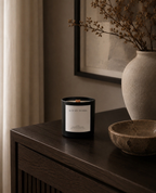 A black glass candle jar with a white label reading 'AUTUMN WOODS' by Luceria Candle Co. sits on a dark wooden console table in a styled interior setting with a ceramic vase and bowl nearby.