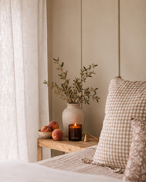 A cozy bedroom corner with a wooden side table holding a beige ceramic vase with dried branches, a lit amber glass candle, and a small bowl of peaches, next to a beige gingham pillow on a bed. Persimmon and Peach Candle by Luceria Candle Co.
