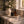 A styled countertop scene by a window with a black candle labeled LUCERIA, an amber bottle, and a brass diffuser on a marble tray, beside a ceramic pitcher of white flowers in a real interior setting.