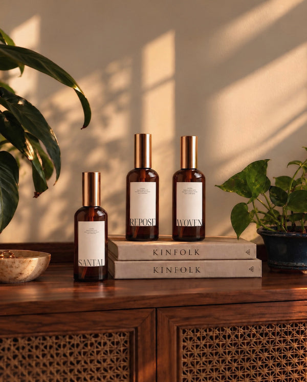 Three amber glass LUCERIA bottles with gold caps and white labels are arranged on stacked books on a wooden cabinet, with potted plants and warm sunlight casting shadows in the background.
