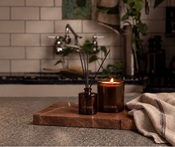 A reed diffuser and a lit amber candle on a wooden tray, placed on a countertop in a bathroom setting with a tiled wall and sink in the background. The products are branded LUCERIA.