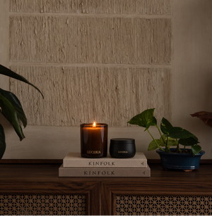 A lit amber glass candle and a small black jar labeled LUCERIA are placed on stacked KINFOLK books on a wooden cabinet, with potted plants on both sides against a textured beige wall.