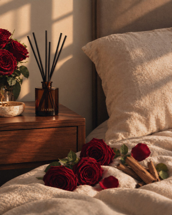 A bedroom scene with a wooden bedside table, a Luceria amber reed diffuser bottle with black reeds, and several deep red roses placed on the bed and in a vase. Warm sunlight casts shadows across the beige bedding and pillow.
