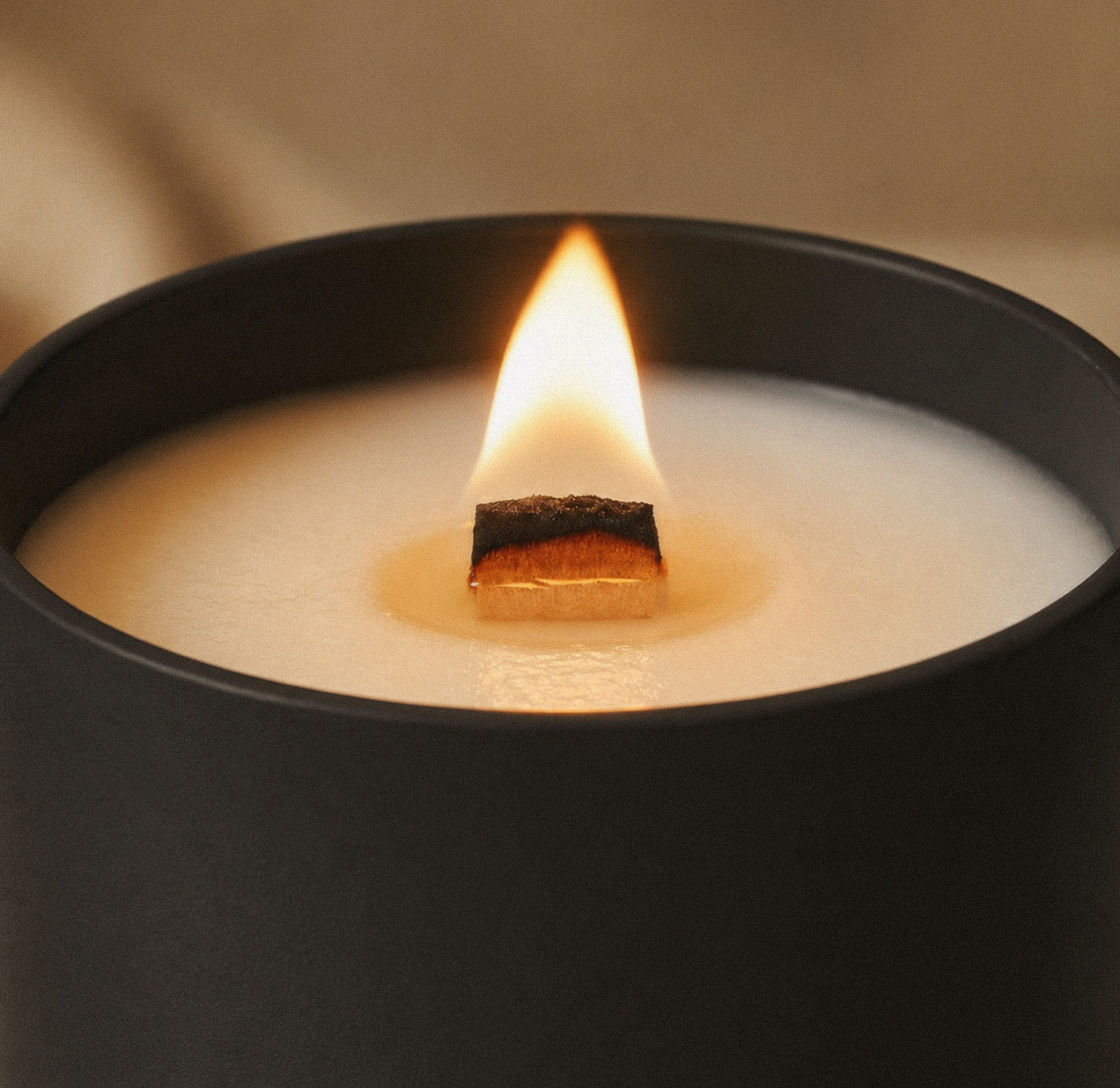 A close-up of a Luceria black candle jar with a single lit wood wick, showing a glowing flame and creamy white wax against a warm beige background.