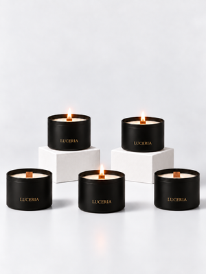 Five black Luceria scented candles with gold lettering are arranged on white pedestals against a clean white background. Three candles are lit, showing a warm flame and cream-colored wax.