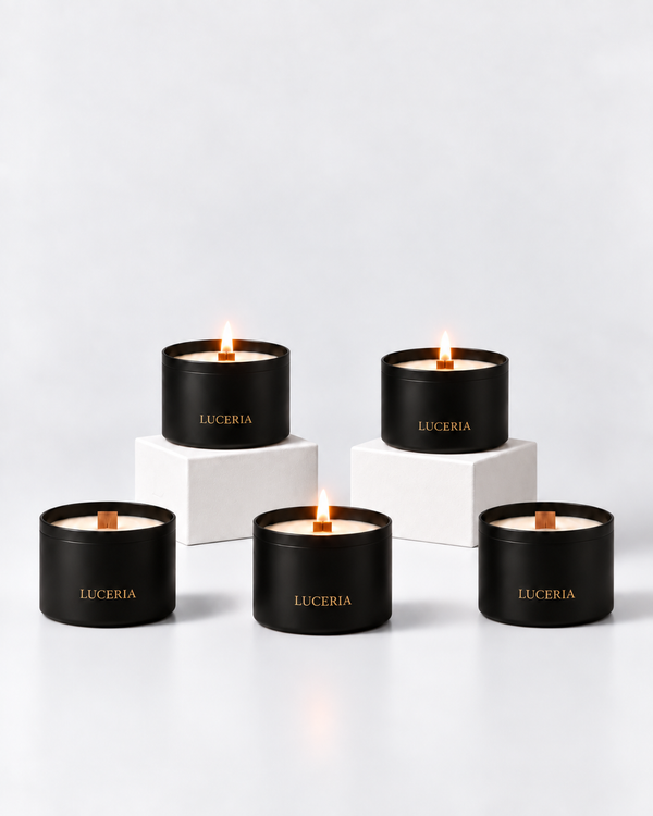 Five black Luceria scented candles with gold lettering are arranged on white pedestals against a clean white background. Three candles are lit, showing a warm flame and cream-colored wax.