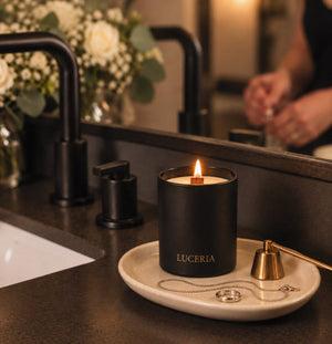 A black Luceria candle with a lit flame sits on a small beige tray on a dark bathroom countertop, with a faucet, mirror, and blurred hands in the background.