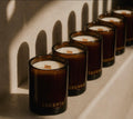 A row of amber glass scented candles with white wax and wooden wicks, branded LUCERIA, arranged diagonally on a light stone surface with strong shadow patterns. Luceria Candle Co. Digital Gift Card.