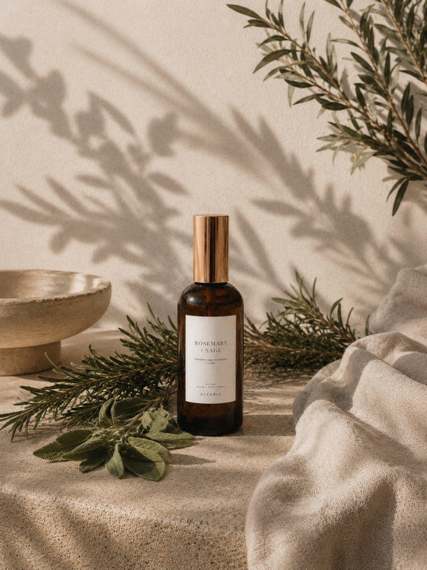 Luceria Rosemary and Sage room spray in an amber glass bottle styled with fresh rosemary, sage leaves, pine sprigs, and neutral linen.