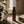 An amber glass bottle with a black cap, labeled 'LUCERA', sits on a stone ledge in warm sunlight next to a folded towel, with a curtain and plant leaves in the foreground. Terra Lemongrass and Patchouli Scented Room and Linen Spray by Luceria Candle Co.