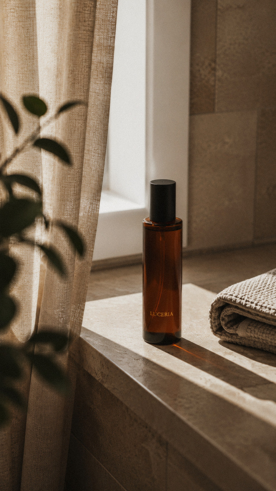 An amber glass bottle with a black cap, labeled 'LUCERA', sits on a stone ledge in warm sunlight next to a folded towel, with a curtain and plant leaves in the foreground. Terra Lemongrass and Patchouli Scented Room and Linen Spray by Luceria Candle Co.