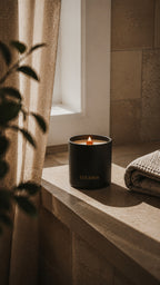 A black cylindrical candle jar with a lit flame sits on a stone windowsill in warm natural light, with beige curtains, a folded towel, and plant leaves in the foreground. The candle label shows the brand LUCERIA. Terra Lemongrass and Patchouli Scented Candle with wood wick by Luceria Candle Co.