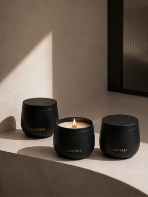 Three matte black LUCERIA candles in rounded glass jars, with the center candle lit and casting a warm glow, arranged on a beige stone ledge in soft sunlight. The Ritual Collection by Luceria Candle Co.
