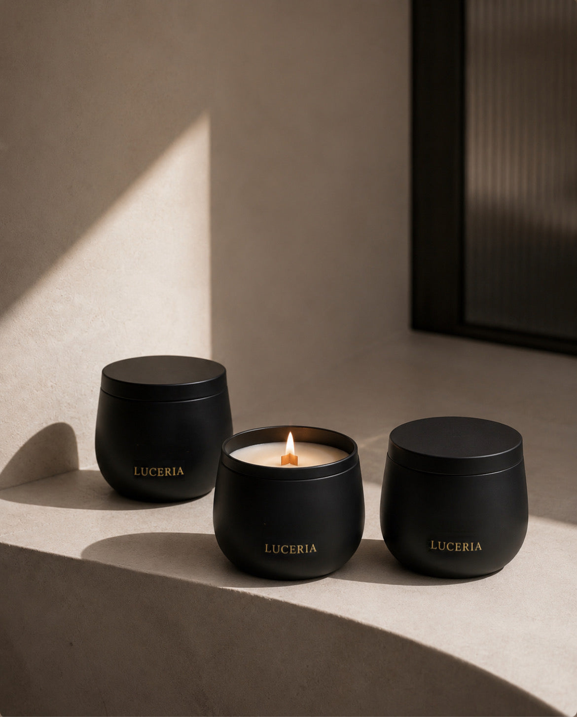 Three matte black LUCERIA candles in rounded glass jars, with the center candle lit and casting a warm glow, arranged on a beige stone ledge in soft sunlight. The Ritual Collection by Luceria Candle Co.