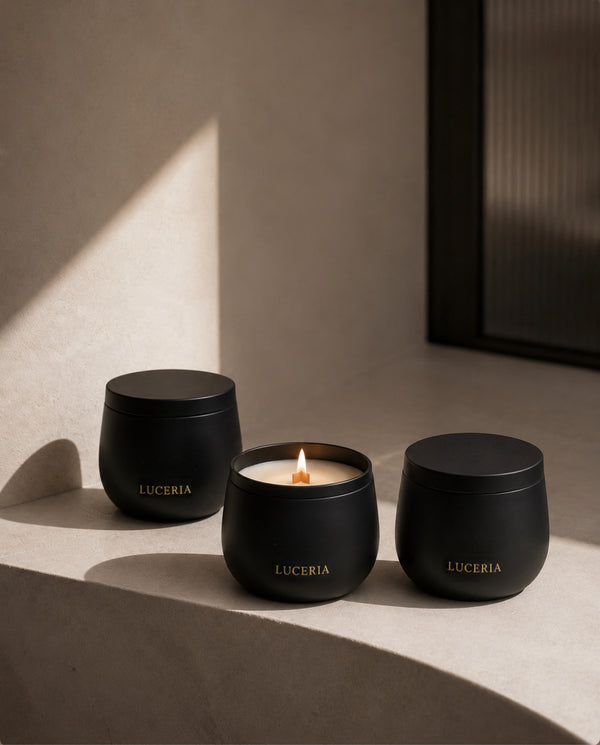 Three matte black LUCERIA candles in rounded glass jars, with the center candle lit and casting a warm glow, arranged on a beige stone ledge in soft sunlight. The Ritual Collection by Luceria Candle Co.