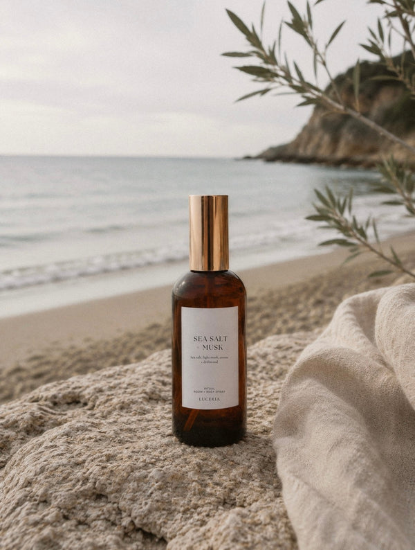 Luceria Sea Salt and Musk room spray in an amber glass bottle styled on a sandy beach with coastal grasses, linen, and ocean shoreline.