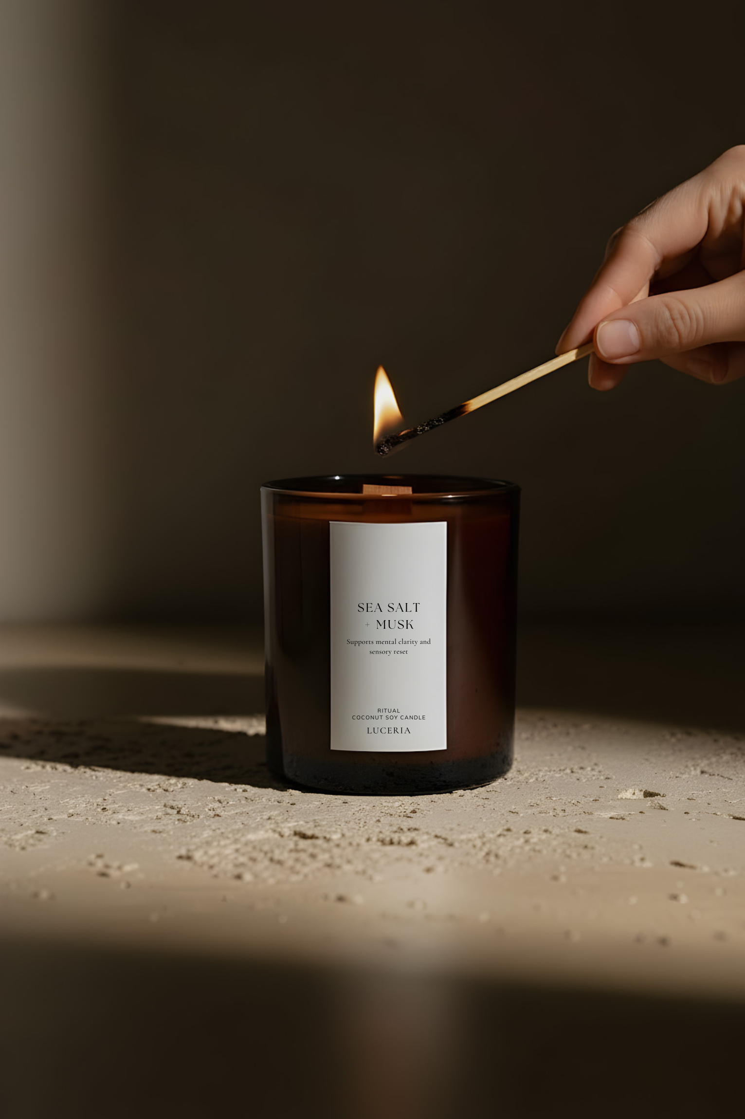 Hand lighting a candle labeled 'Sea salt + musk' candle with a match against a dark background 