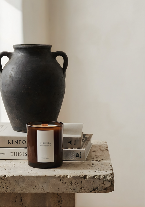 Candle with a label on a stone surface next to a black vase and books.