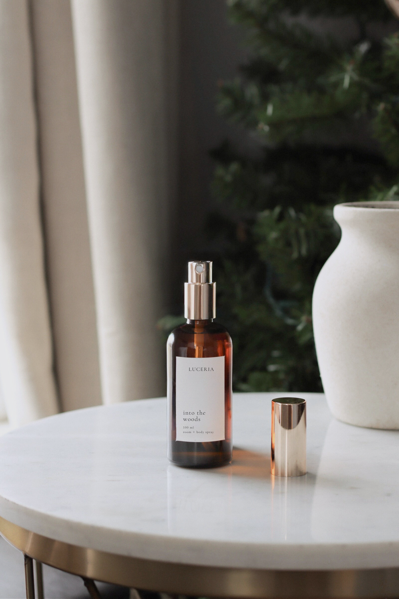 Bottle of "Into the Woods" room and body spray  on a marble table with a plant and vase in the background