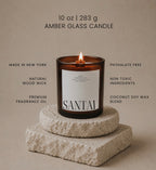 A lit Luceria amber glass candle labeled 'SANTAL' sits on stacked stone blocks against a warm beige background, with text listing product features such as 10 oz size, made in New York, natural wood wick, and coconut soy wax blend.