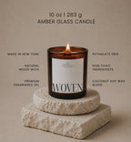 A lit Luceria amber glass candle with a white label reading 'WOVEN', displayed on stacked stone blocks against a warm beige background. Text around the candle highlights product claims such as made in New York, natural wood wick, premium fragrance oil, phthalate free, non toxic ingredients, and coconut soy wax blend.