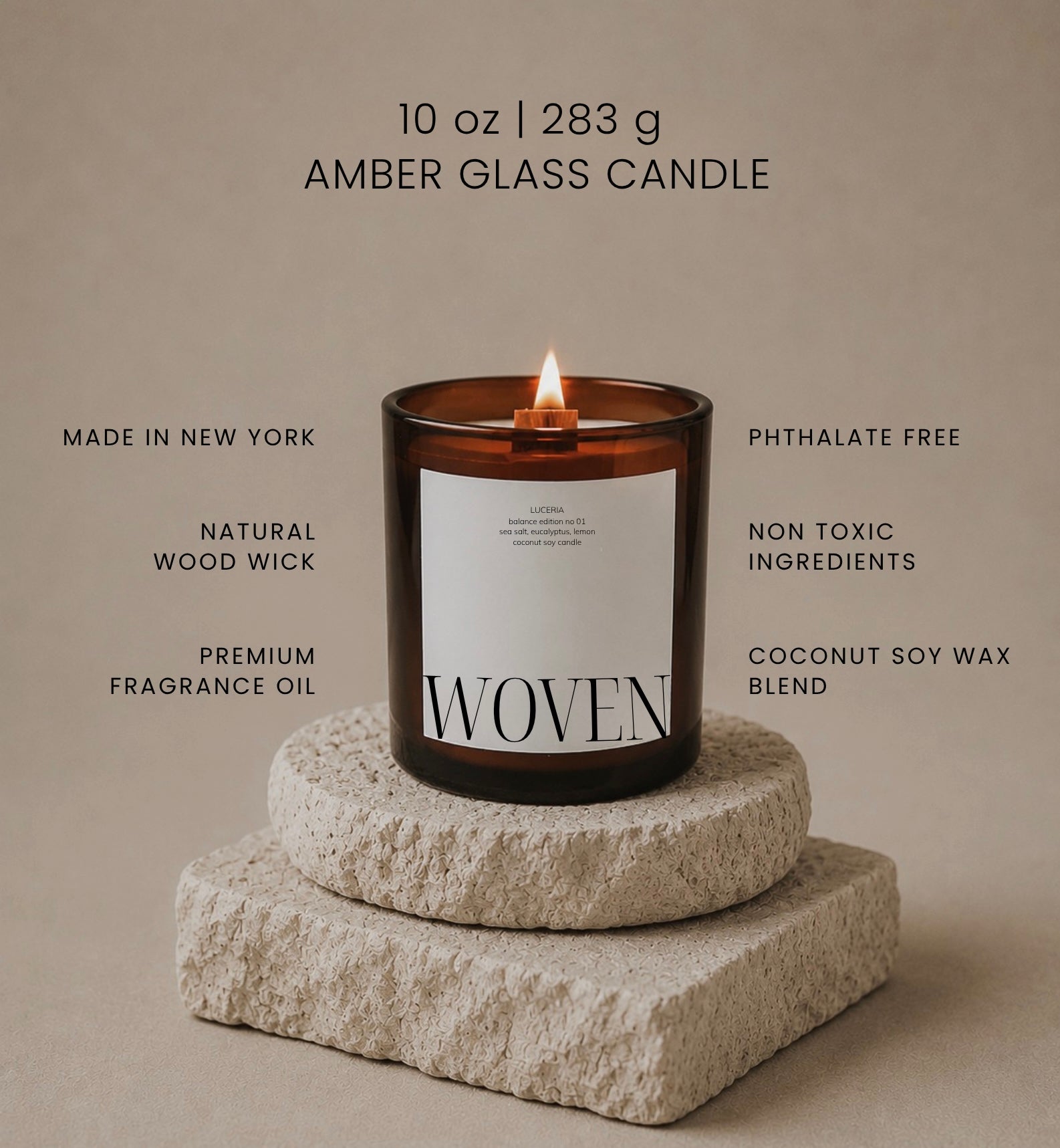 A lit Luceria amber glass candle with a white label reading 'WOVEN', displayed on stacked stone blocks against a warm beige background. Text around the candle highlights product claims such as made in New York, natural wood wick, premium fragrance oil, phthalate free, non toxic ingredients, and coconut soy wax blend.