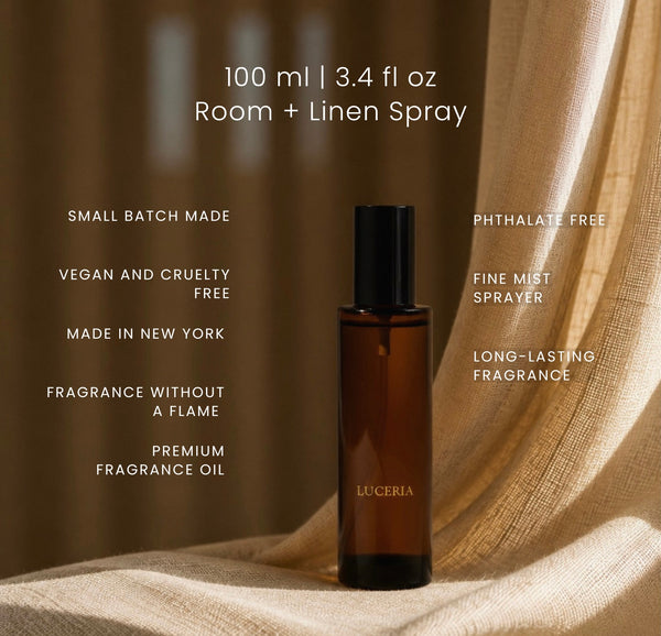 A 100 ml amber glass fragrance oil bottle with a black cap, labeled LUCERIA, set against draped beige linen fabric. White text on the image lists product features such as 'Small batch made', 'Vegan and cruelty free', and 'Phthalate free'.