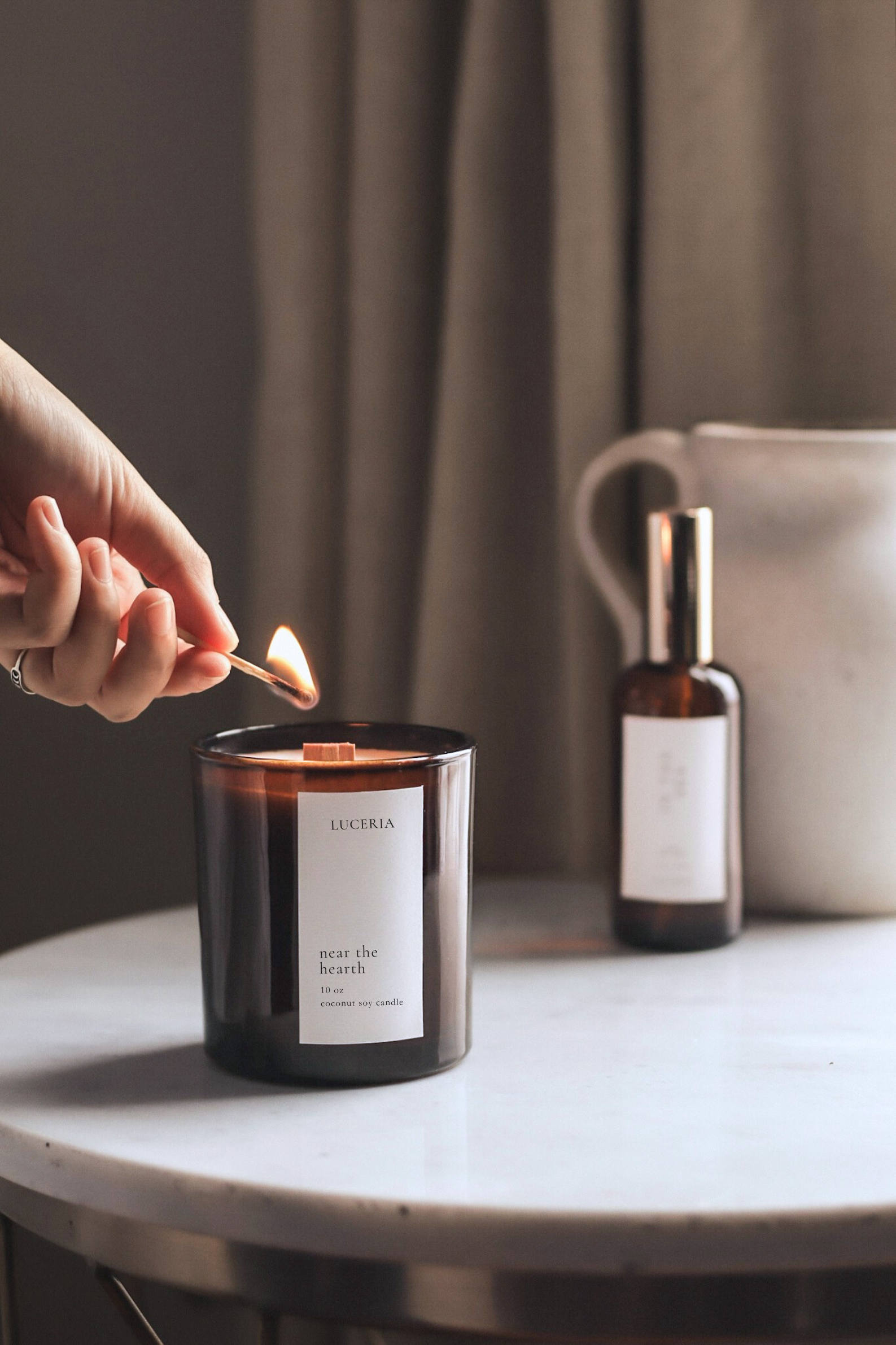 Person lighting Luceria candle with a match on a table next to a bottle and a bag.