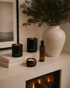 Three Luceria candles and a matching bottle arranged on a fireplace mantel beside a large textured cream vase with dried greenery. The candles are in matte black holders, with one larger candle and two smaller candles, including one lit candle in front.