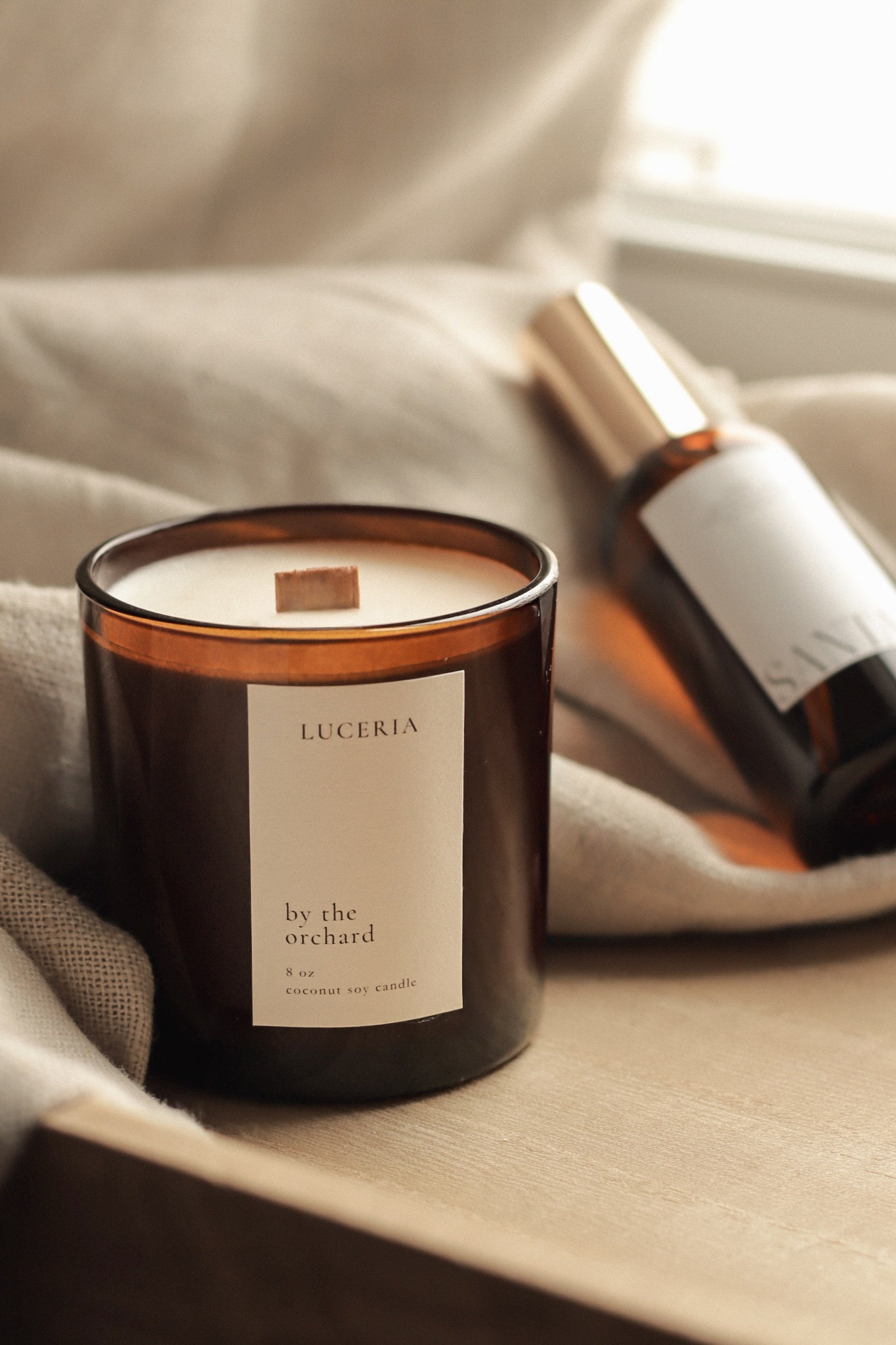 Candle labeled 'Luceria by the orchard' on a soft surface with a bottle in the background.
