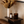 A warm, styled interior scene showing a dark amber glass 'LUCERIA' candle jar and a matching amber 'LUCERIA' bottle with a gold cap on a dark wooden side table. A white vase with dried flowers sits behind them against a beige wall, with part of a brown leather chair visible in the foreground.