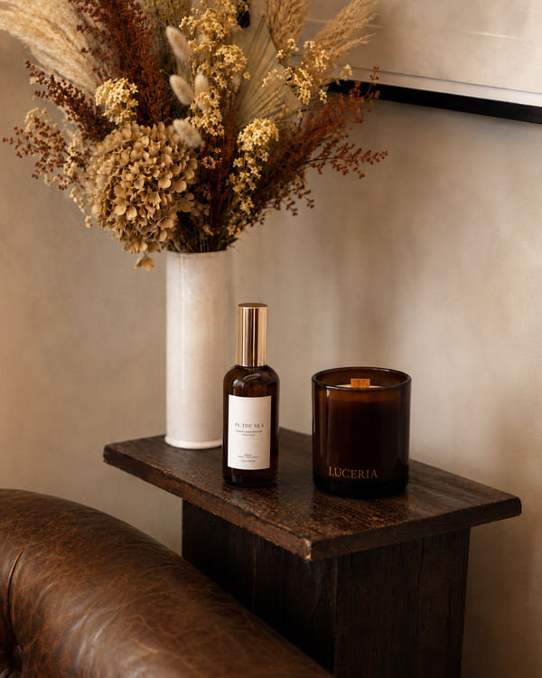 A warm, styled interior scene showing a dark amber glass 'LUCERIA' candle jar and a matching amber 'LUCERIA' bottle with a gold cap on a dark wooden side table. A white vase with dried flowers sits behind them against a beige wall, with part of a brown leather chair visible in the foreground.