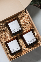 Three small candles in a gift box labeled 'Santal', 'Woven', and 'Repose' in a holiday box with shredded paper.
