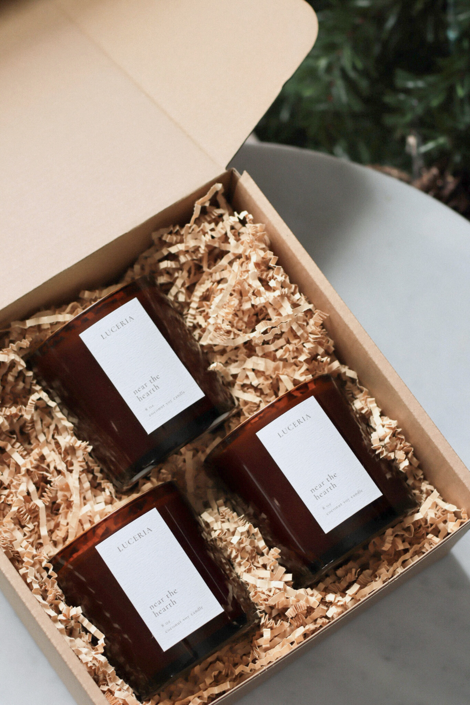 Three candles in a gift box with shredded paper, on a light surface with a blurred green background.