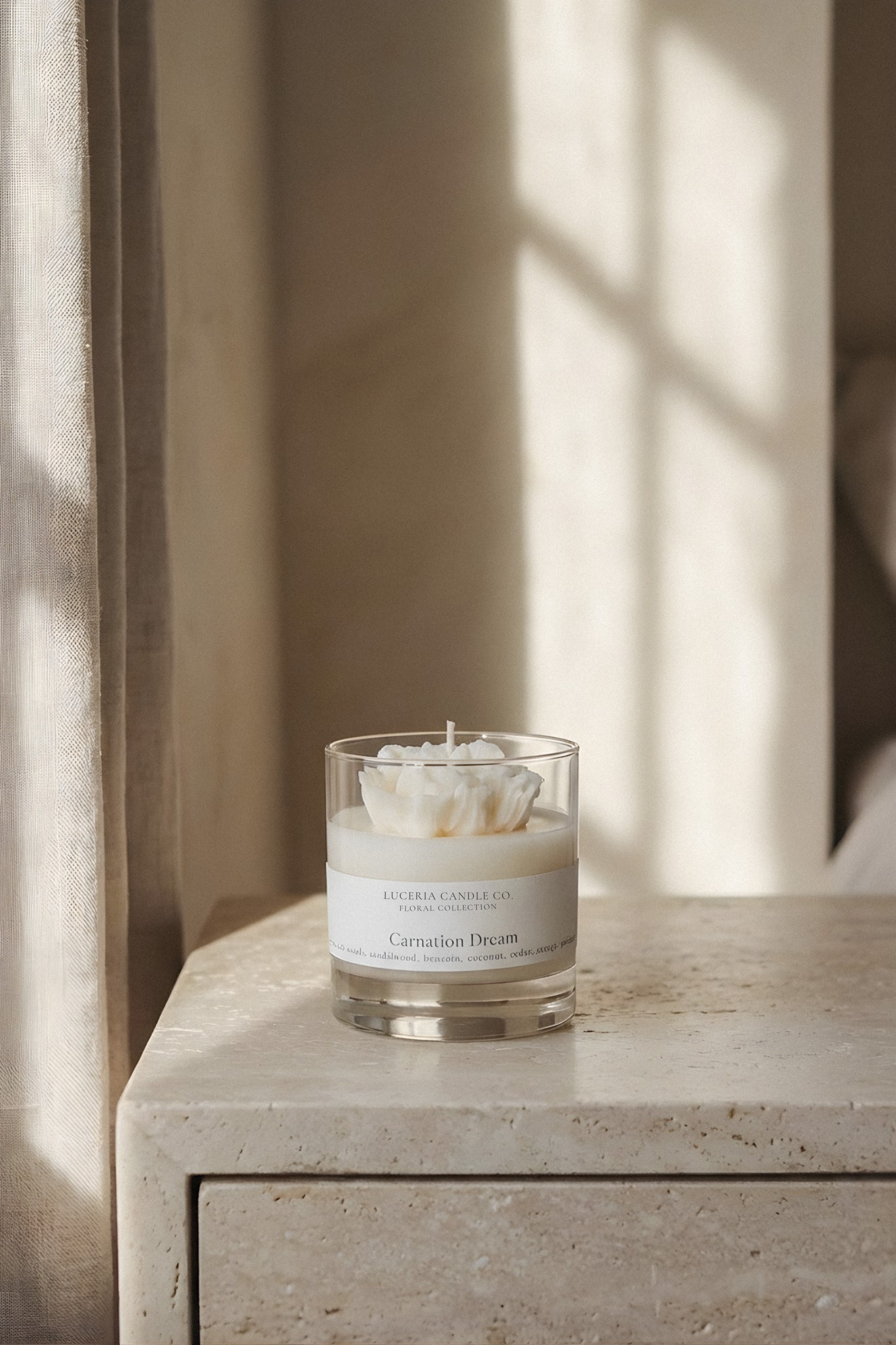Luceria Carnation candle in a glass container on a stone surface with sunlight casting shadows.