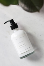Hand lotion bottle labeled 'Cleanse' on a light surface with a plant in the background