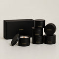 Six black cylindrical tins with gold 'LUCERIA' branding are arranged on a light gray studio background, including one open tin showing white wax and a wooden wick, plus a matching black gift box.
