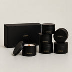 Six black cylindrical tins with gold 'LUCERIA' branding are arranged on a light gray studio background, including one open tin showing white wax and a wooden wick, plus a matching black gift box.
