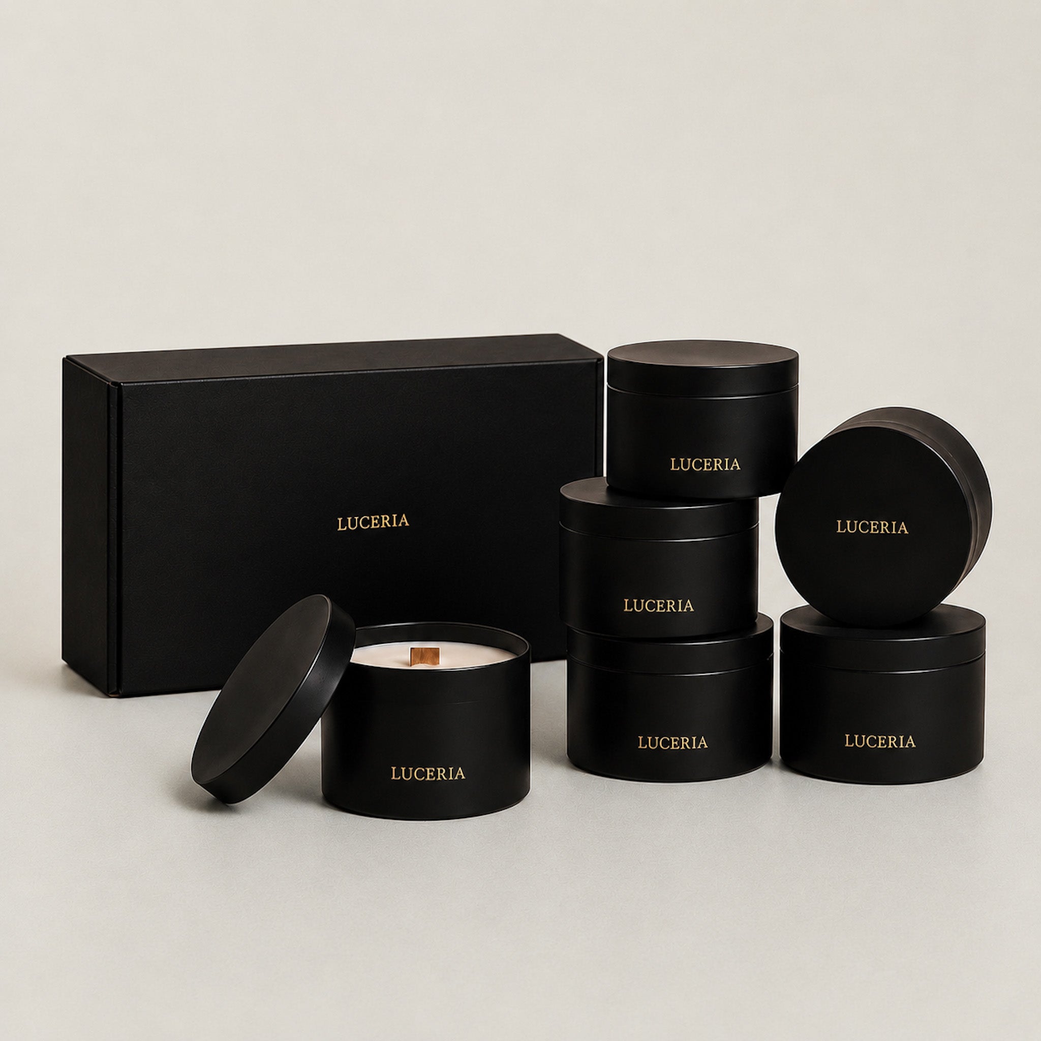 Six black cylindrical tins with gold 'LUCERIA' branding are arranged on a light gray studio background, including one open tin showing white wax and a wooden wick, plus a matching black gift box.