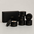 Six black cylindrical tins with gold 'LUCERIA' branding are arranged on a light gray studio background, including one open tin showing white wax and a wooden wick, plus a matching black gift box. The Earthen Collection Set, 7 oz matte black candle tins by Luceria Candle Co.