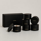 Six black cylindrical tins with gold 'LUCERIA' branding are arranged on a light gray studio background, including one open tin showing white wax and a wooden wick, plus a matching black gift box. The Earthen Collection Set, 7 oz matte black candle tins by Luceria Candle Co.