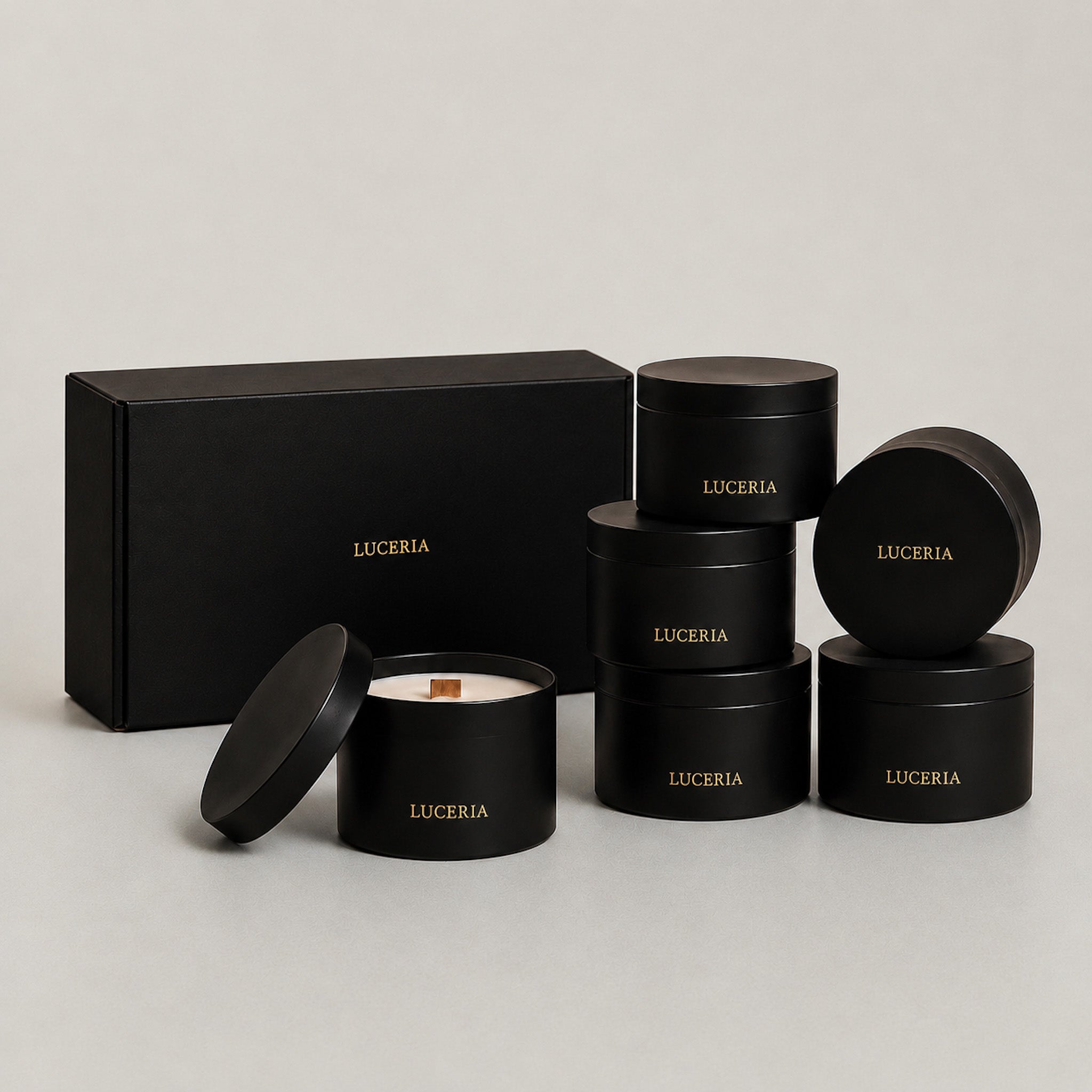 Six black cylindrical tins with gold 'LUCERIA' branding are arranged on a light gray studio background, including one open tin showing white wax and a wooden wick, plus a matching black gift box. The Earthen Collection Set, 7 oz matte black candle tins by Luceria Candle Co.