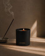 Ember Palo Santo + Sandalwood Candle by Luceria burning in a matte black glass vessel beside smoking incense on a beige surface.