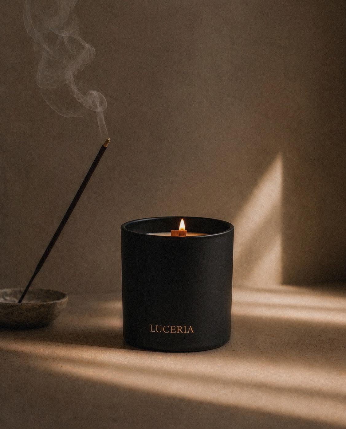 A matte black candle jar with the brand name 'LUCERIA' in gold lettering, lit with a small flame. It is styled on a textured beige surface with dramatic warm sunlight casting shadows, next to a smoking incense stick in a bowl.