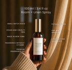 A hand in a beige knit sweater holds a bottle of Luceria Lavender Cedar Room + Linen Spray against a dark brown background with draped beige fabric. White text on the image lists product features and scent details.