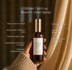 A hand in a beige knit sweater holds a bottle of Luceria Redwoods and Moss Room + Linen Spray against a dark brown background with draped beige fabric. White text on the image lists product features and volume.