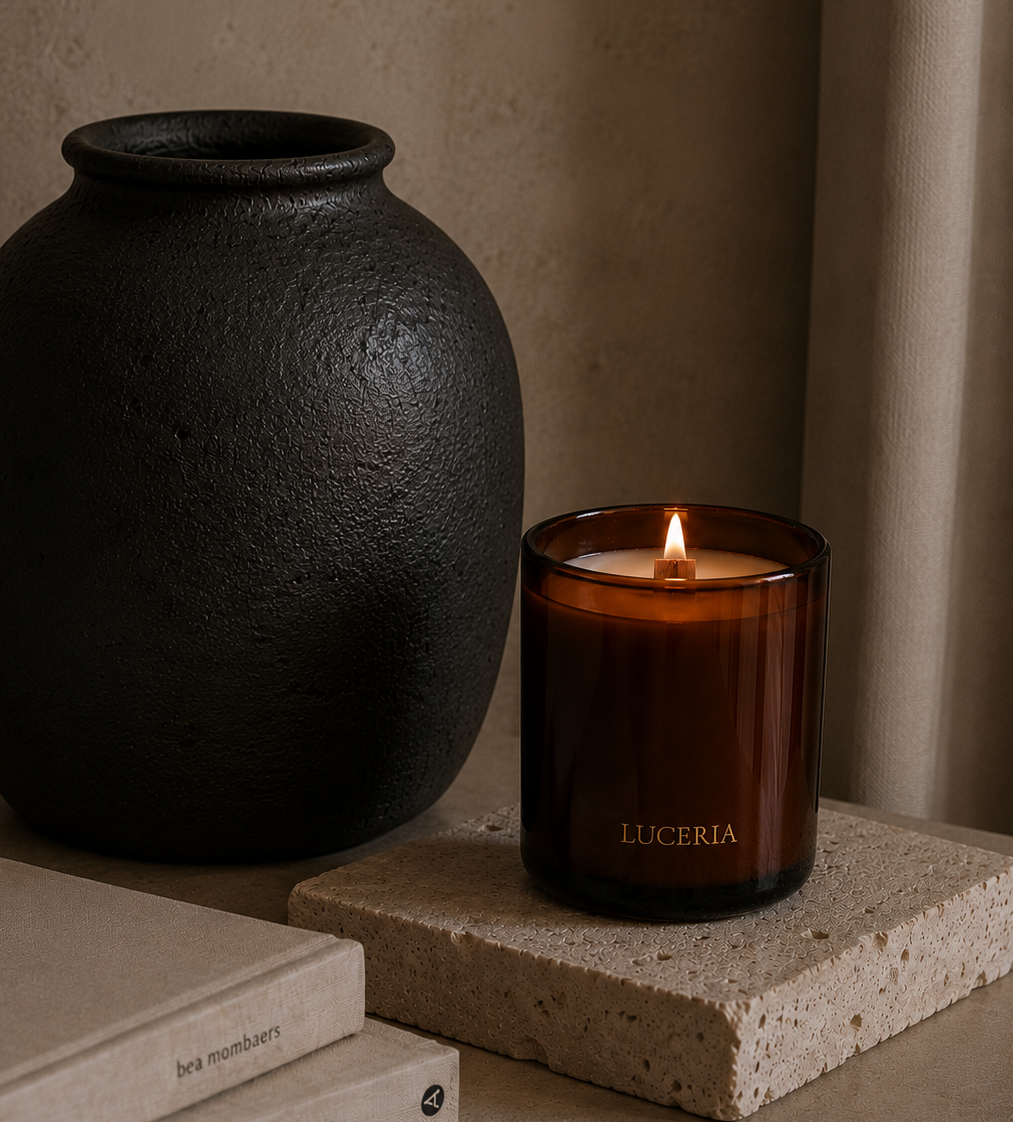 An amber glass candle jar with a lit flame and the brand name LUCERIA, styled on a stone slab next to a large textured black vase and neutral-toned books.
