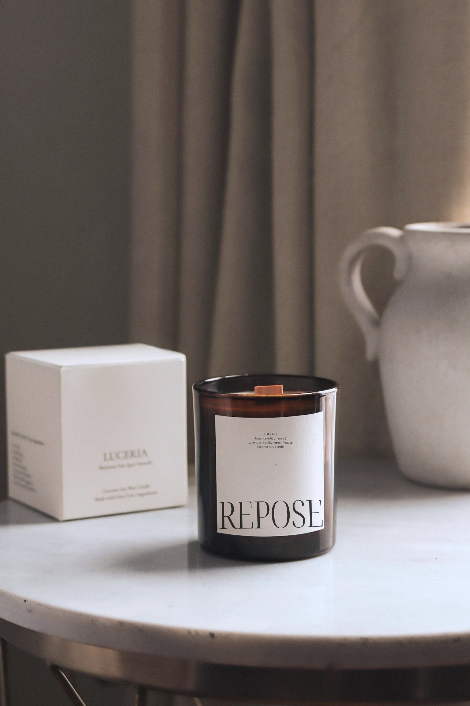 Candle labeled 'Repose' on a table with a box and ceramic pitcher in the background