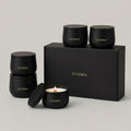 A set of matte black LUCERIA candles and matching boxes on a light gray studio background. The set includes two stacked black jars, one open jar with a lit candle, and a large rectangular gift box, all with gold LUCERIA branding. The Ritual Collection Set, 7 oz matte black candle tins by Luceria Candle Co.