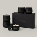A set of matte black LUCERIA candles and matching boxes on a light gray studio background. The set includes two stacked black jars, one open jar with a lit candle, and a large rectangular gift box, all with gold LUCERIA branding. The Ritual Collection Set, 7 oz matte black candle tins by Luceria Candle Co.